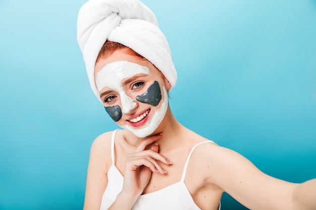 Leuke vrouw met gezichtsmasker selfie met oprechte glimlach te nemen. Studio shot van blanke meisje poseren tijdens huidverzorgingsroutine.