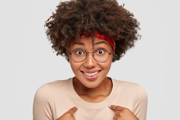 Leuke zwarte vrouw lacht vrolijk, voelt trots op haar daden, vraagt iets, is verbaasd dat ze werd uitgekozen, heeft een krullend kapsel, modellen tegen een witte muur, overweldigd door positieve emoties