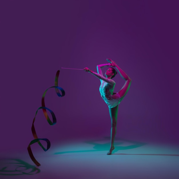 Levendig. Jonge vrouwelijke atleet, ritmische gymnastiek artiest dansen, trainen met tape geïsoleerd op paarse studio achtergrond in neonlicht. Mooi meisje oefenen met apparatuur. Genade in uitvoering