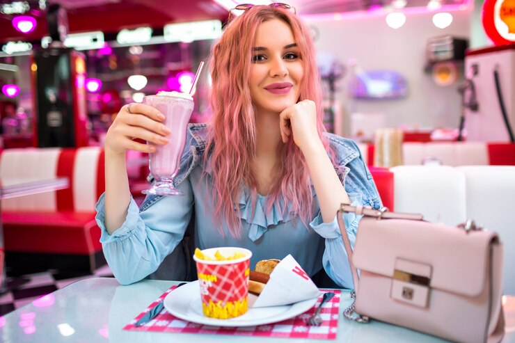 Gratis Foto levensstijl binnenbeeld van stijlvolle jonge mooie vrouw met golvende ongebruikelijke roze haren en natuurlijke make-up, schattige blauwe jurk en spijkerjasje dragen, geniet van haar smakelijke amerikaanse diner.
