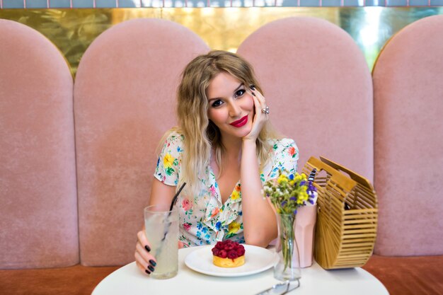 Levensstijl schattig beeld van mooie blonde vrouw poseren, zitten en genieten van haar maaltijd, kijken op camera, elegante bloemenjurk en lichte make-up, frambozencake eten