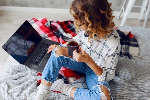 Levensstijlbeeld, vrouw die koffie drinkt en computer gebruikt, warme sokken en trendy jeans draagt. Zittend op bed. Vroege morgen. Bovenaanzicht.