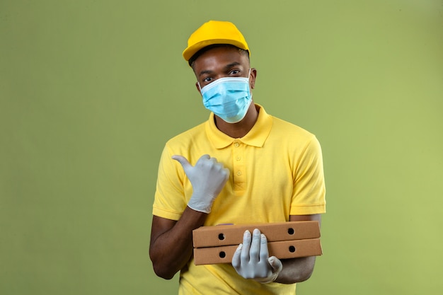 Levering Afro-Amerikaanse man in geel poloshirt en pet dragen van medische beschermend masker houden pizzadozen met glimlach op gezicht wijzend naar de kant met duim staande op groen