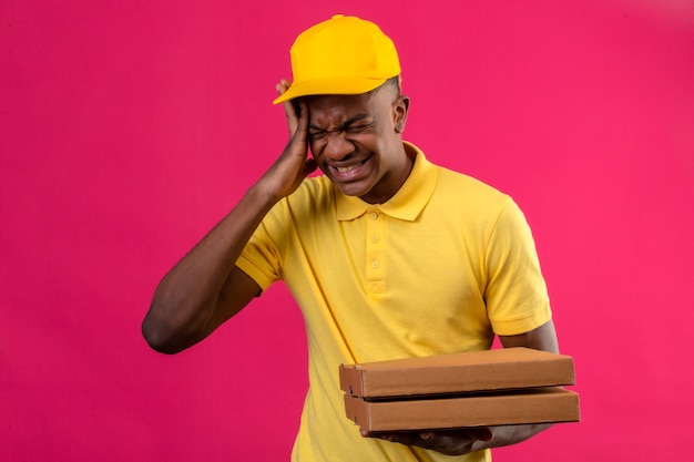 Levering Afro-Amerikaanse man in geel poloshirt en pet permanent met hand op het hoofd onwel lijdt aan sterke hoofdpijn op roze