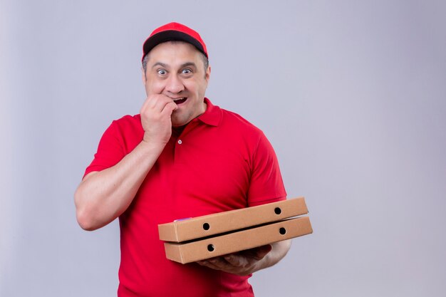Levering man in rood uniform en pet houden pizzadozen op zoek gestrest en nerveus met hand op mond nagels bijten staande op wit
