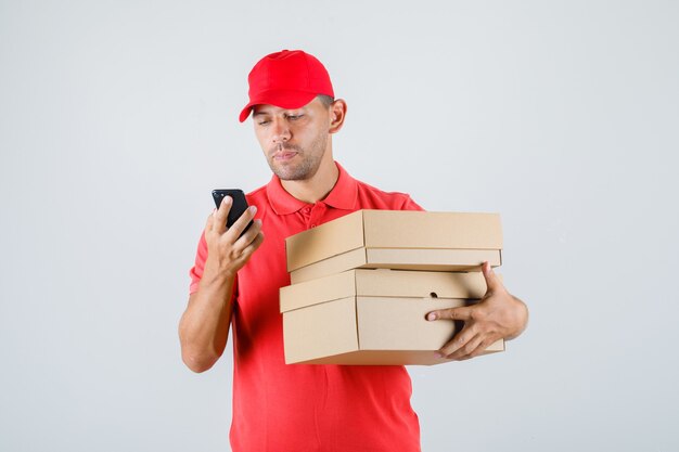 Levering man in rood uniform kartonnen dozen te houden tijdens het gebruik van smartphone