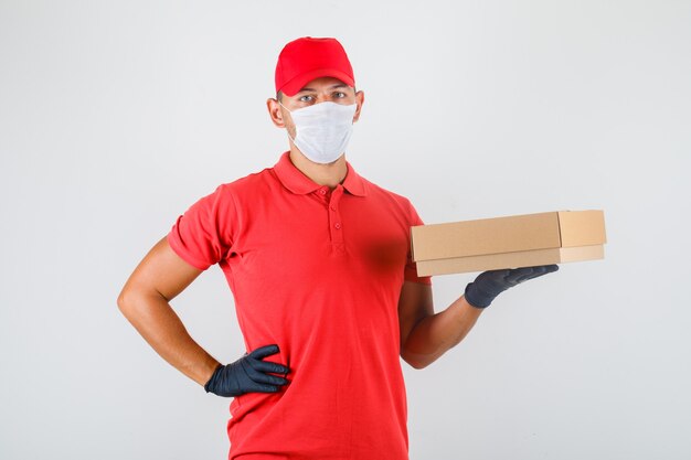 Levering man met kartonnen doos met hand op taille in rood uniform, medisch masker, handschoenen vooraanzicht.