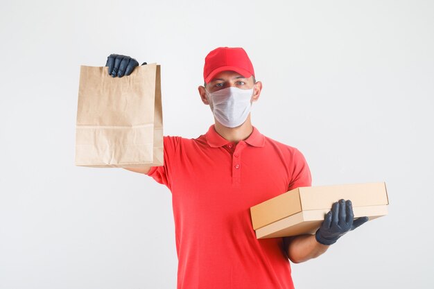 Levering man met papieren zak en kartonnen doos in rood uniform, medisch masker, handschoenen vooraanzicht.