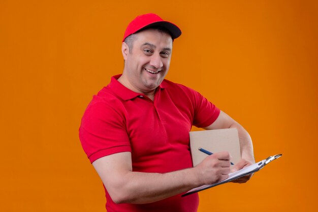 Levering man met rode uniform en pet met doos pakket en klembord met pen glimlachend vrolijk staande ruimte