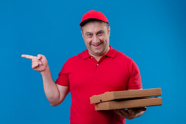 Levering man met rode uniform en pet met pizzadozen lachend met blij gezicht wijzend met wijsvinger naar de zijkant staande over blauwe ruimte