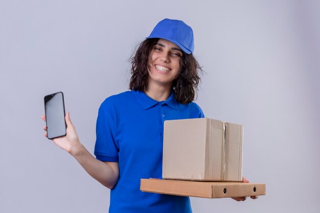 Levering meisje in blauw uniform en pet met pizzadozen en doos pakket met mobiele telefoon glimlachend vrolijk staande op wit