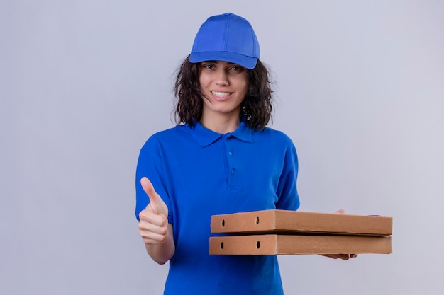 Levering meisje in blauw uniform en pet met pizzadozen met glimlach op gezicht duimen opdagen staande op wit