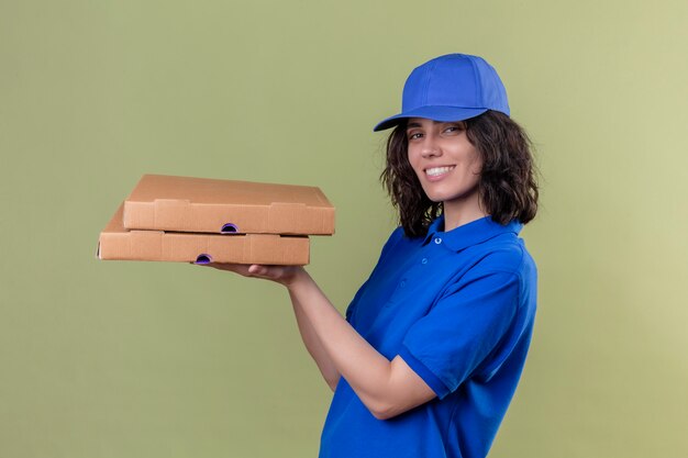 Levering meisje in blauw uniform en pet met pizzadozen op zoek blij positief en gelukkig lachend vrolijk staande op geïsoleerde groen