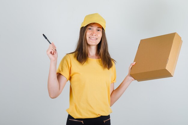 Levering vrouw in t-shirt, broek, pet met kartonnen doos en pen en ziet er vrolijk uit