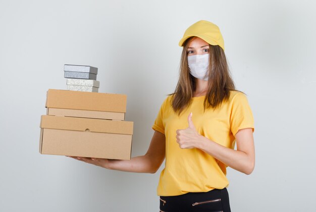 Levering vrouw met dozen met duim omhoog in t-shirt, broek en pet, masker en op zoek vrolijk