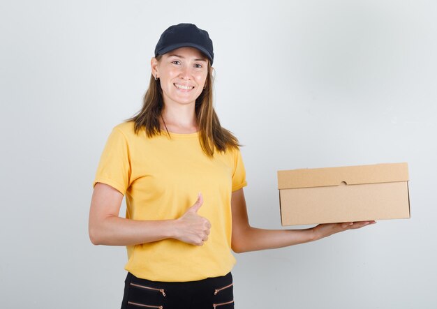 Levering vrouw met kartonnen doos met duim omhoog in t-shirt, broek en pet en op zoek vrolijk