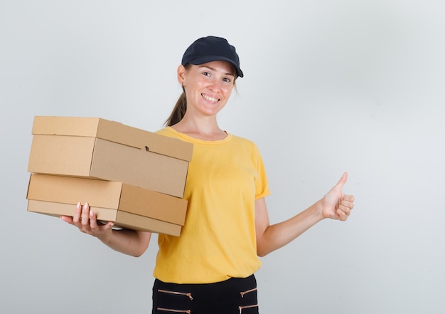 Levering vrouw met kartonnen dozen met duim omhoog in t-shirt, broek, pet en op zoek vrolijk