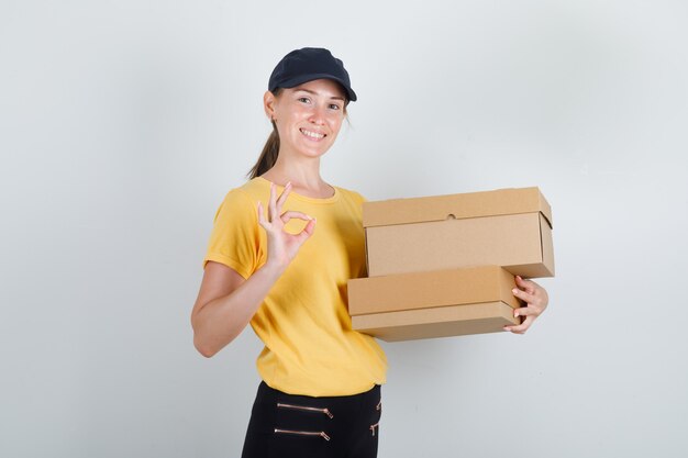 Gratis foto levering vrouw met kartonnen dozen met ok teken in t-shirt, broek, pet en op zoek vrolijk.