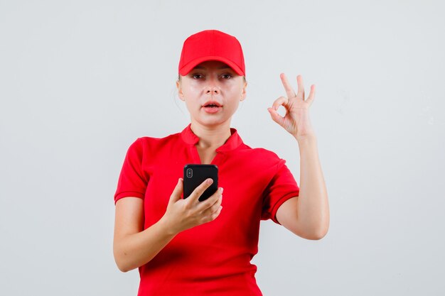 Levering vrouw met mobiele telefoon met ok gebaar in rood t-shirt en pet en op zoek tevreden