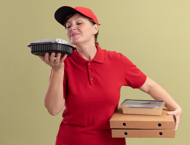 Levering vrouw van middelbare leeftijd in rood uniform en pet met pizzadozen en voedselpakketten gelukkig en positief inademen aangenaam aroma staande over groene muur