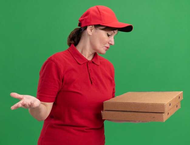 Levering vrouw van middelbare leeftijd in rood uniform en pet met pizzadozen kijken naar hen verward staande over groene muur