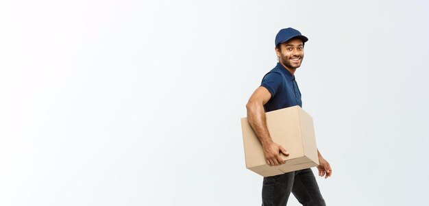Leveringsconcept Portret van een gelukkige Afro-Amerikaanse bezorger in blauwe doek die loopt om een doospakket naar de klant te sturen. Geïsoleerd op een grijze studioachtergrond