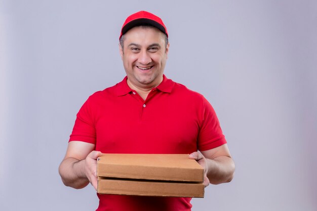 Leveringsmens die rode eenvormig en GLB-de pizzadozen van de holdings glimlachen vriendelijk met gelukkig gezicht over geïsoleerde witte muur dragen