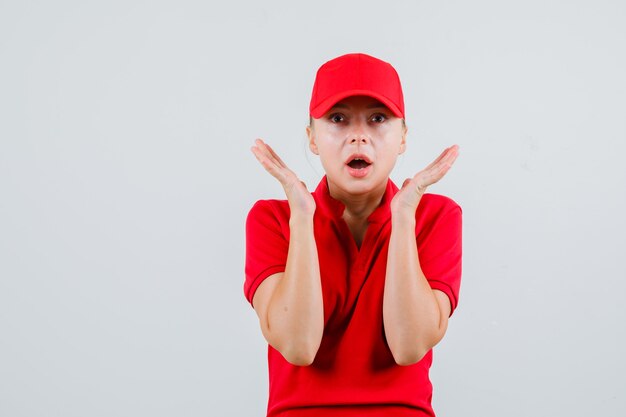 Leveringsvrouw in rood t-shirt en pet die opgeheven handpalmen dichtbij gezicht houdt en verbaasd kijkt