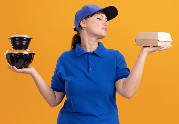 Leveringsvrouw van middelbare leeftijd in blauw uniform en pet met voedselpakketten die zelfverzekerd en gelukkig staande over oranje muur kijken