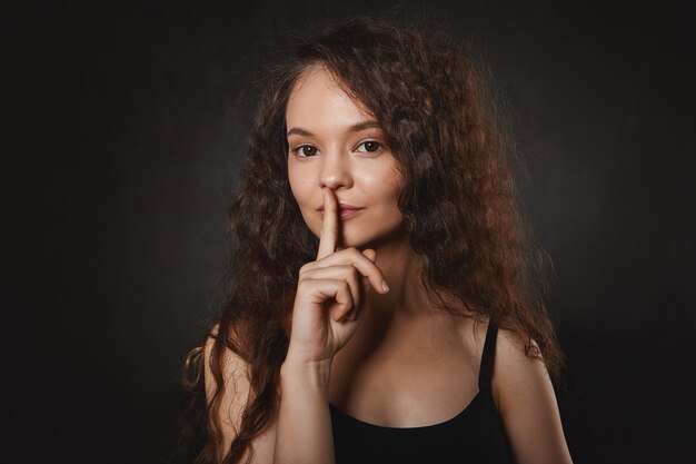 Lichaamstaal. Portret van mooie charmante jonge Europese vrouw met zwarte ogen en mysterieuze glimlach, wijsvinger vasthoudend aan haar mond, met groot geheim, Shh zeggen