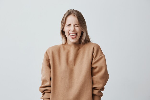 Lichaamstaal. Weerzinwekkende gestreste mooie vrouw met steil blond haar poseert, fronst haar gezicht, sluit ogen en steekt haar tong uit, wat een afkeer van iets toont