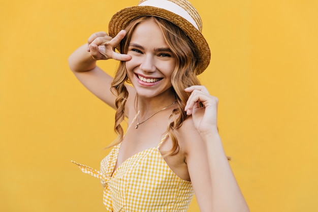 Lichtbruine krullende vrouw in hoed het glimlachen. Binnenopname van geïnspireerd vrouwelijk model draagt geruite gele kleding.