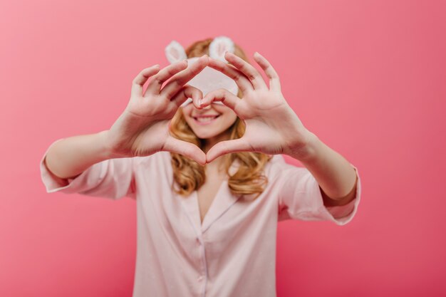 Lief meisje dat in oogmasker hartteken toont. Binnenportret van glimlachende krullende vrouw in zijden pyjama die op roze muur met haar handen op voorgrond wordt geïsoleerd.