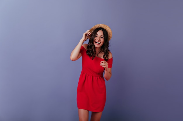 Lief meisje met bruin haar lachen op paarse muur. indoor foto van vrolijke brunette dame draagt rode jurk en zomerhoed.