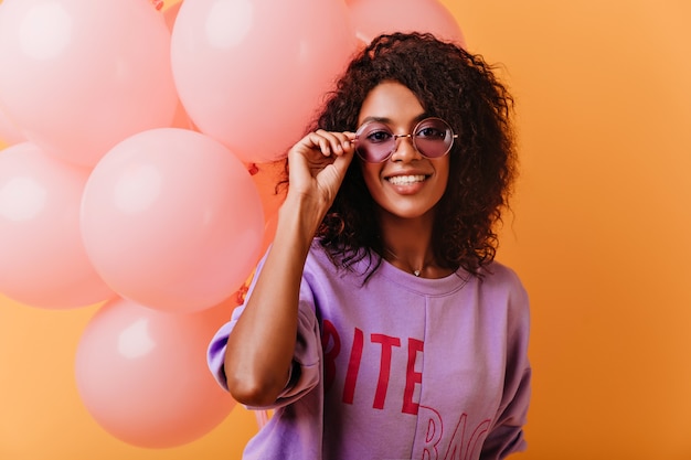 Lief vrouwelijk model in glazen en paars shirt poseren op feestje. Enthousiaste zwarte jonge vrouw met plezier tijdens feestelijk.