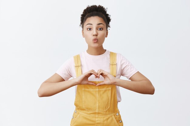 Liefde, zorg en relatie concept. Portret van charmante tedere en liefdevolle Afro-Amerikaanse vrouw in stijlvolle gele overall, lippen vouwen in mwah en hart teken tonen over borst, kus willen