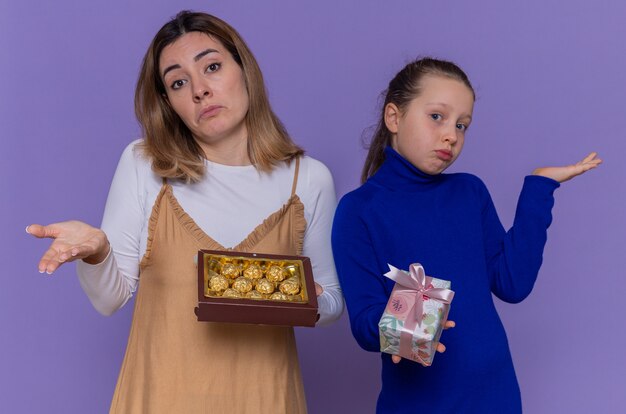 Liefdevolle moeder met doos chocolaatjes en dochter bedrijf aanwezig op zoek verward armen opheffen vieren internationale vrouwendag staande over paarse muur