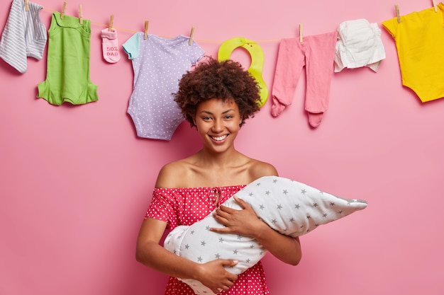 Liefdevolle vrolijke moeder houdt haar kleine twee maanden oude zoon gewikkeld in een deken, speelt met baby, verantwoordelijke jonge moeder, geniet van momenten van moederschap, staat tegen touw met babykleertjes