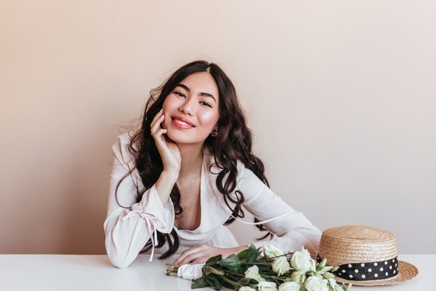 Lieve Aziatische vrouw die met krullend haar bij camera glimlacht. Studio shot van charmant Japans model met witte bloemen.