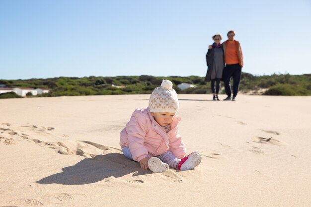 Lieve baby die roze jasje en hoed draagt, buiten op zand speelt