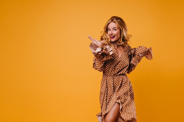 Lieve blonde meisje poseren met verbaasde glimlach. Indoor foto van geweldige Europese vrouw dansen op gele muur.