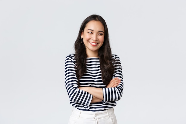 Lifestyle, beauty en mode, mensen emoties concept. Vrolijk schattig en verlegen Aziatisch meisje met kruisarmen borst bescheiden pose, wegkijkend als lachend en dom lachend, op een witte achtergrond staan