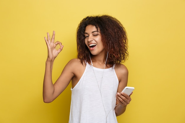 Lifestyle Concept - Portret van mooie Afrikaanse Amerikaanse vrouw blij muziek aan de mobiele telefoon te luisteren. Gele pastel studio achtergrond. Ruimte kopiëren.