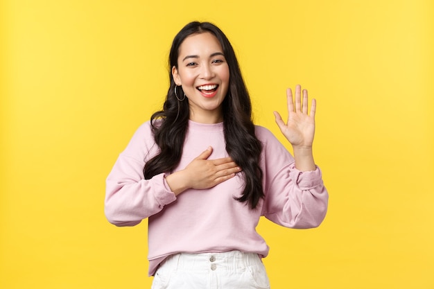 Lifestyle, emoties en advertentieconcept. Eerlijke en oprechte gelukkige glimlachende aziatische vrouw belooft de waarheid te vertellen, zweer op haar hart en steekt één hand op, staande gele achtergrond.
