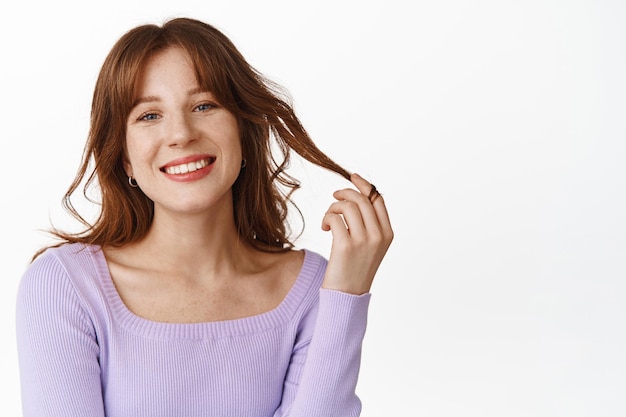 Lifestyle en mode concept. Mooie en stijlvolle vrouw raakt haar haar aan, nieuw kapsel met pony, ziet er gelukkig en glimlachend uit, draagt een paarse blouse, witte achtergrond