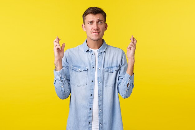 Lifestyle, mensen emoties en zomer vrije tijd concept. Bezorgde blonde knappe man maakte zich zorgen over zijn toekomst, kruiste vingers en bijtende lip als wens, wachtend op belangrijke resultaten, gele achtergrond.