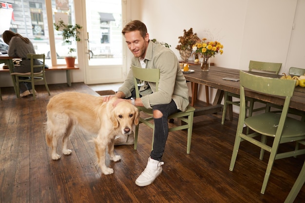 Lifestyle portret van een knappe glimlachende man die in een huisdiervriendelijk café zit met zijn mooie hond