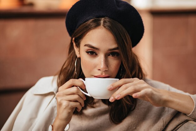 Lifestyle portret van elegante jonge vrouw met donker golvend kapsel, trendy make-up, modieuze beige trui en jas, zittend op café terras en koffie drinken uit witte kop