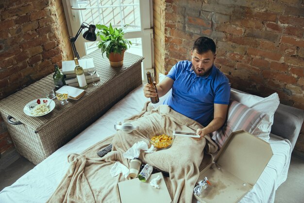 Luie man die in zijn bed woont, omringd door rommelig. Je hoeft niet de deur uit om gelukkig te zijn