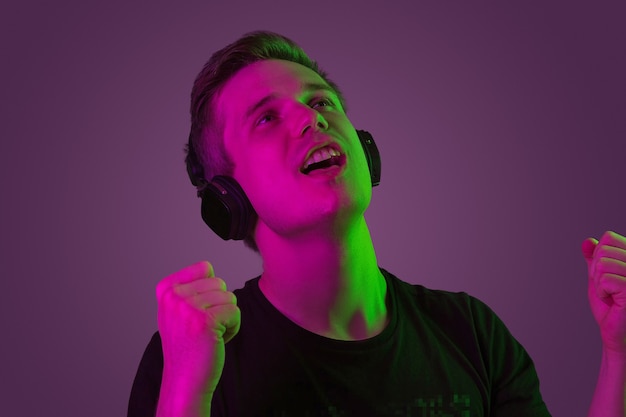 Luister naar muziek, zing. Blanke man portret geïsoleerd op paarse studio achtergrond in neonlicht. Mooi mannelijk model in zwart overhemd. Concept van menselijke emoties, gezichtsuitdrukking, verkoop, advertentie.